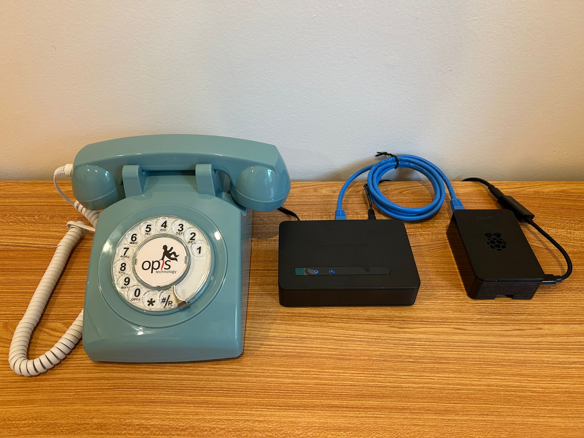 A powder-blue Opis rotary telephone sits to the left of two small black electronic devices connected by a blue ethernet cable on a wooden surface. The devices are the Grandstream HT812 analog telephone adapter and a Raspberry Pi 4 in a black case — the complete hardware required for the Calls from the Field listening station.
