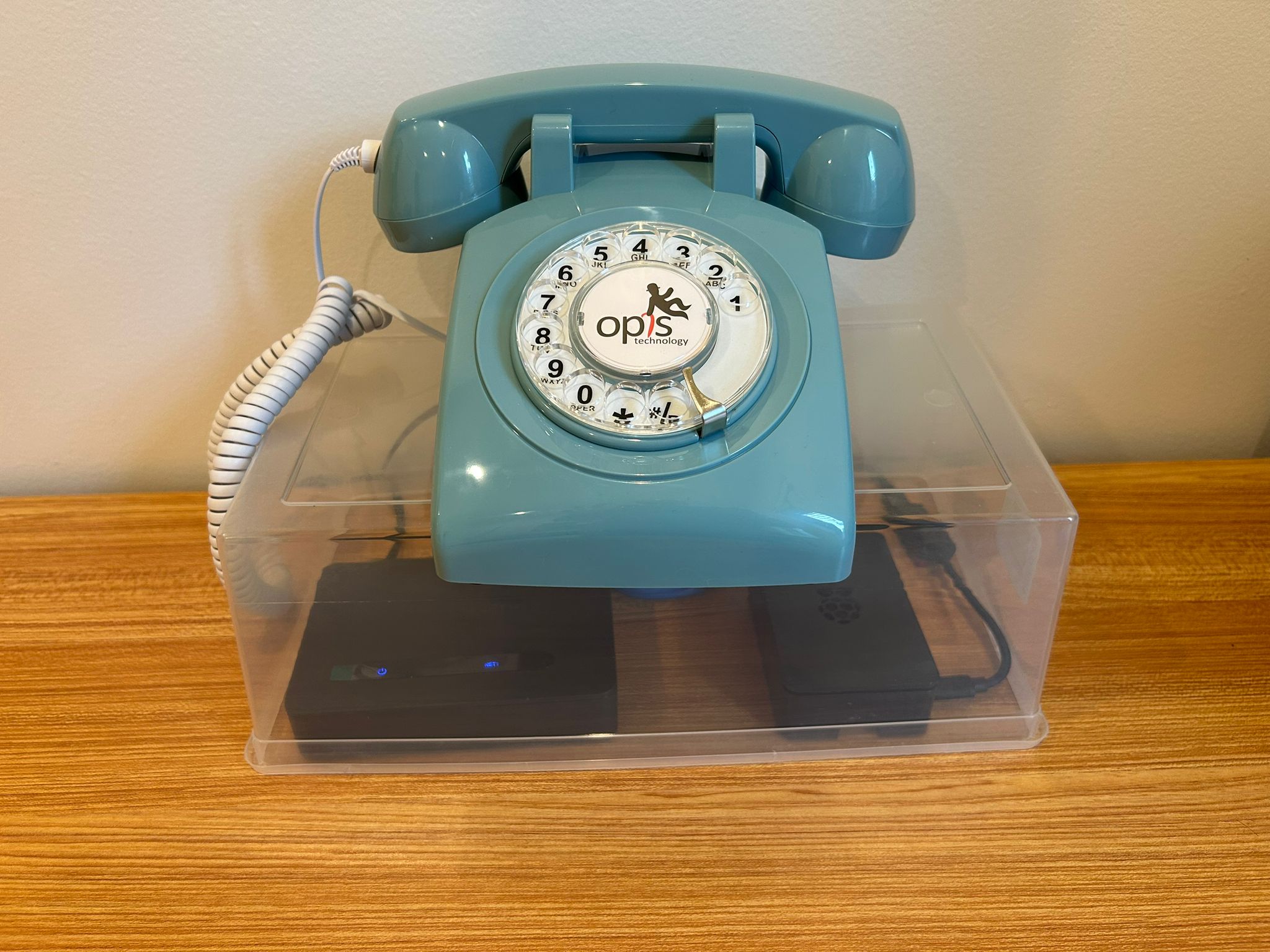 A powder-blue rotary telephone sits on a clear acrylic box on a wooden surface. Through the transparent walls of the box, the Grandstream HT812 adapter and Raspberry Pi 4 are visible underneath, showing the full system in its assembled display configuration.