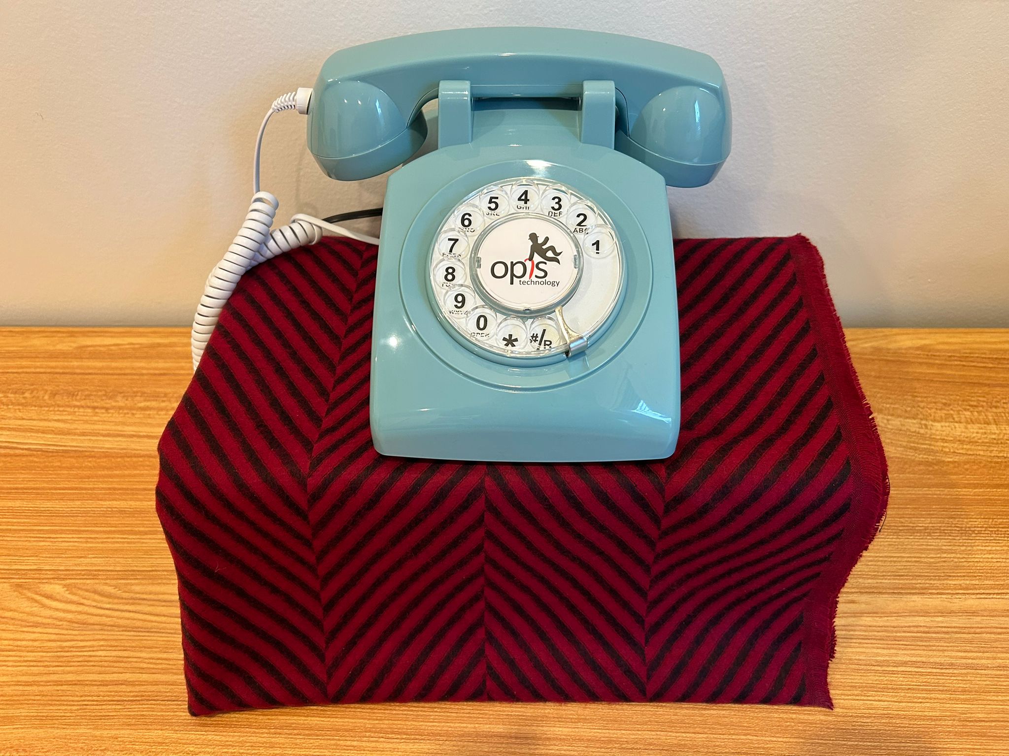 A powder-blue rotary telephone sits on a folded red textile with a chevron pattern on a wooden surface. The adapter and Raspberry Pi are hidden beneath the fabric, leaving only the telephone visible — an alternative display configuration that emphasizes the phone as the primary object of interaction.