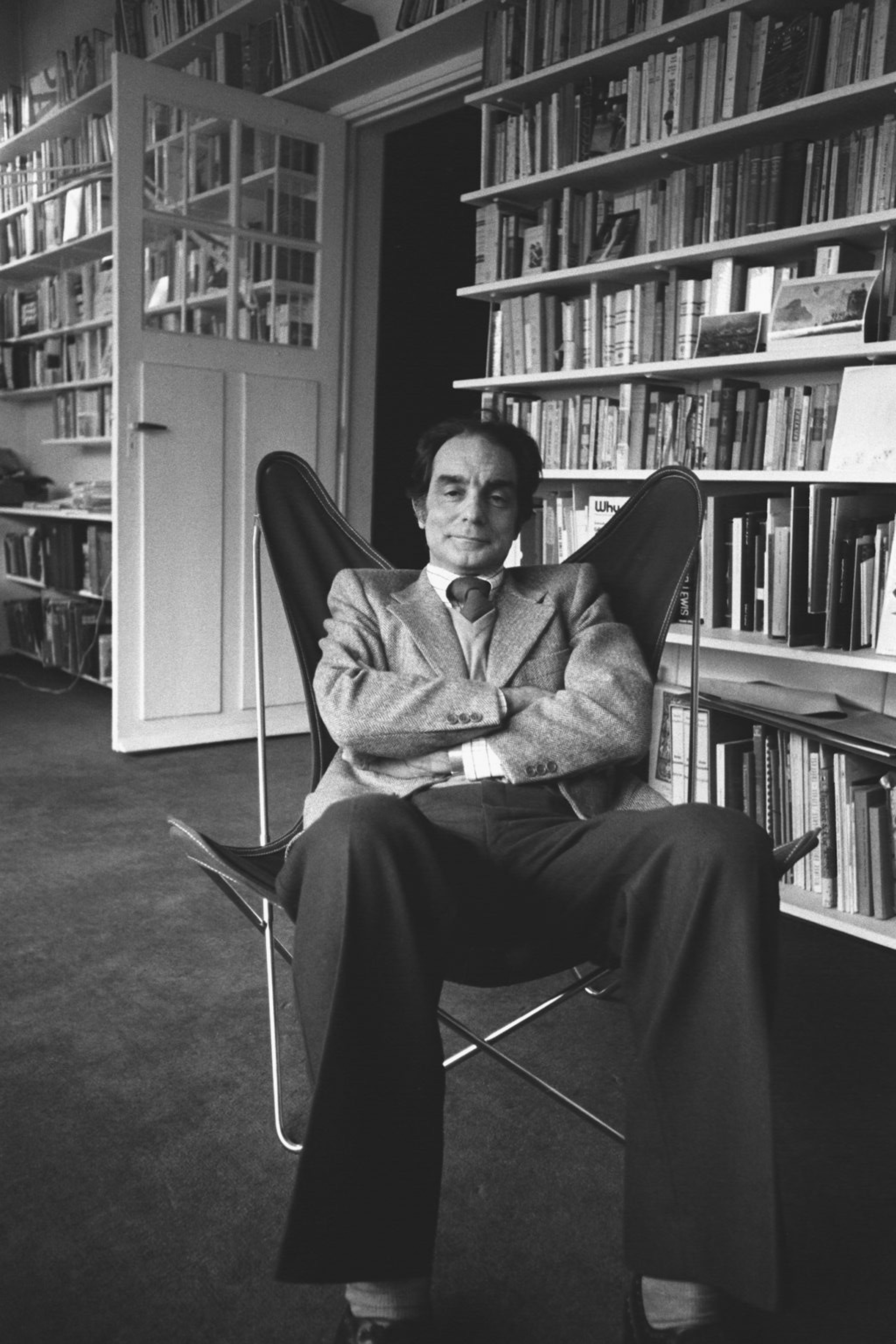 Black-and-white photograph of Italo Calvino seated in a low chair with his arms crossed, surrounded by tall bookshelves in a study or library.